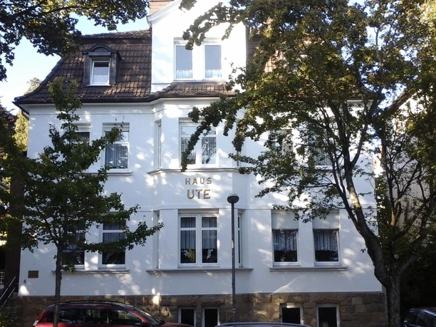 021 Ein altes, weißes Gebäude mit dem Schriftzug "Haus Ute". Umgeben von Bäumen und einer ruhigen Straße.