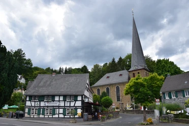 Ensemble Volberg <p>Historisches Fachwerkhaus neben einer alten Kirche mit spitzem Turm unter bewölktem Himmel.</p>