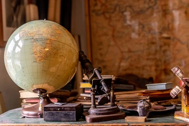 Heimatmuseum Ein antiker Globus, ein Mikroskop und Bücher sind auf einem Holztisch vor einer alten Weltkarte arrangiert.