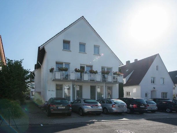 Ein modernes, weißes Gebäude mit Balkonen und mehreren Autos davor. Der Himmel ist klar und die Sonne scheint hell.