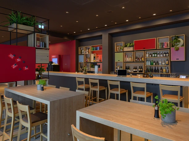 Herford_Restaurant_BarTeutoburger-Wald-Tourismus-InterCity-Hotel.jpg Moderne Restaurantbar mit Holztischen, hellen Holzstühlen, offener Regale mit Flaschen und Pflanzen.