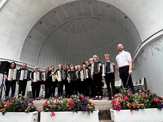 Bad Driburg_Akkordeon Konzert_Gräeflicher Park.jpg Akkordeonorchester mit elf Mitgliedern und Dirigent steht vor einer Konzertbühne im Freien.