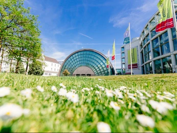 Glasgebäude mit halbrunder Fassade, umgeben von Grünflächen und blühenden Blumen unter blauem Himmel.