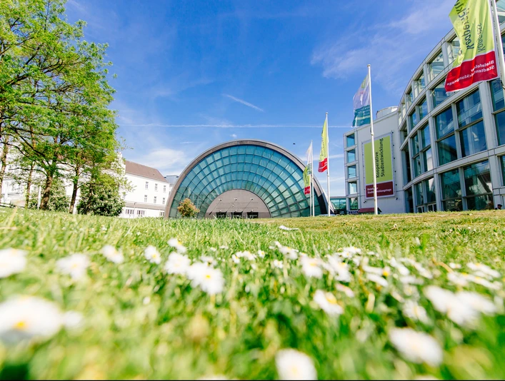 Glasgebäude mit halbrunder Fassade, umgeben von Grünflächen und blühenden Blumen unter blauem Himmel.