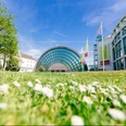 Stadthalle Bielefeld mit Messehalle.jpg Glasgebäude mit halbrunder Fassade, umgeben von Grünflächen und blühenden Blumen unter blauem Himmel.