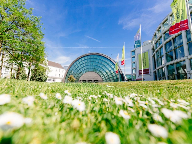 Stadthalle Bielefeld mit Messehalle.jpg Glasgebäude mit halbrunder Fassade, umgeben von Grünflächen und blühenden Blumen unter blauem Himmel.