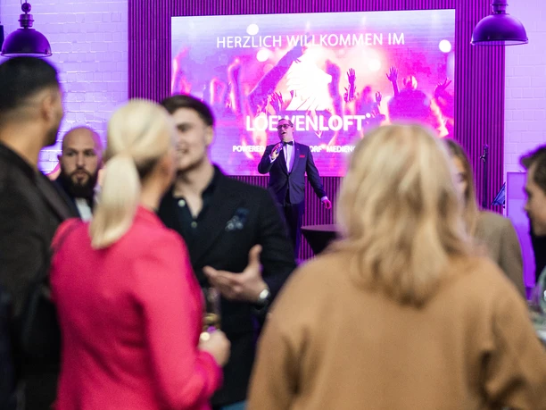 Impressionen Events In the foreground are people at an event, in the background is a man in a black sacko on stage, behind him a screen with the inscription "Welcome to the Löwenloft"