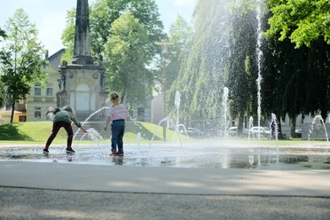 Teutoburger_Wald_Detmold_Kasulke_KaiserWilhelmPl_CCBYSA (3).JPG Kinder spielen an einem Wasserspielplatz vor einem historischen Denkmal, umgeben von Bäumen.