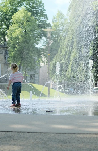 Teutoburger_Wald_Detmold_Kasulke_KaiserWilhelmPl_CCBYSA (3).JPG Kinder spielen an einem Wasserspielplatz vor einem historischen Denkmal, umgeben von Bäumen.