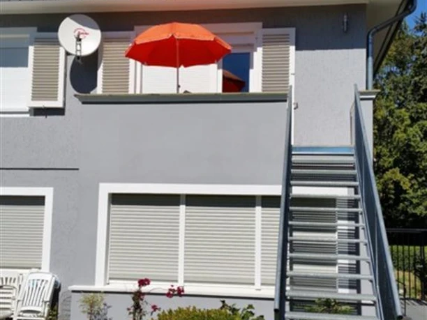 Ein zweigeschossiges Haus mit grauer Fassade und einem roten Sonnenschirm auf dem Balkon. Eine Treppe führt zum Eingang und der Garten ist gepflegt.