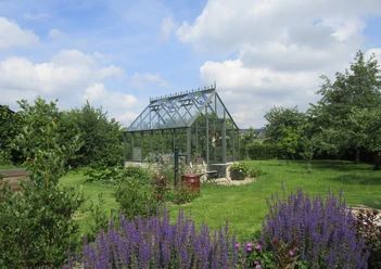 Garten Kottkamp Ein Gewächshaus steht in einem blühenden Garten mit Lavendel im Vordergrund unter einem bewölkten Himmel.