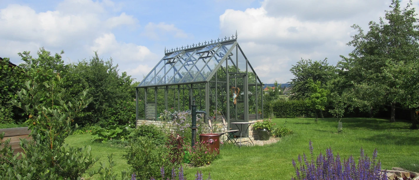 Garten Kottkamp Ein Gewächshaus steht in einem blühenden Garten mit Lavendel im Vordergrund unter einem bewölkten Himmel.