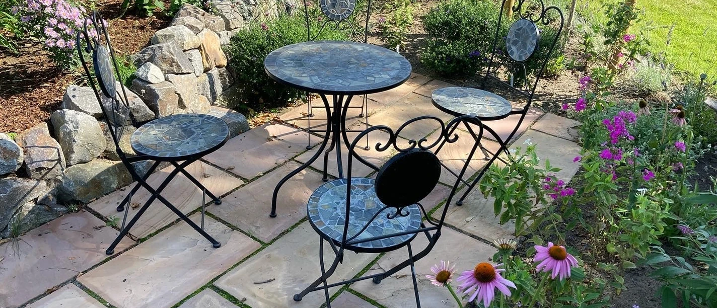 Steintisch und Stühle aus Metall stehen auf einer Terrasse inmitten bunter Blumen im Garten Hartmann.