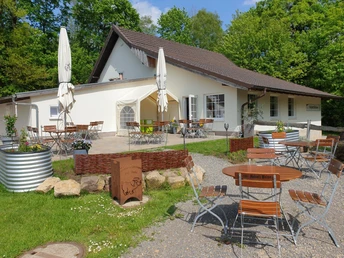 Weißes Landhaus mit braunem Dach, umgeben von Außensitzplätzen aus Holz, umrahmt von grünen Bäumen.