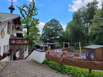 Außenbereich der Waldgaststätte Malepartus, umgeben von Bäumen und rustikalen Tischen auf Pflastersteinen.