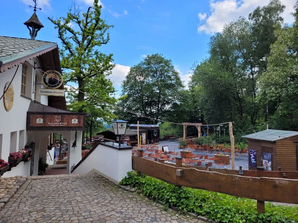 Lienen_Hermannshöhen_Malepartus Waldgaststätte_Anja Schmidt.jpg Außenbereich der Waldgaststätte Malepartus, umgeben von Bäumen und rustikalen Tischen auf Pflastersteinen.