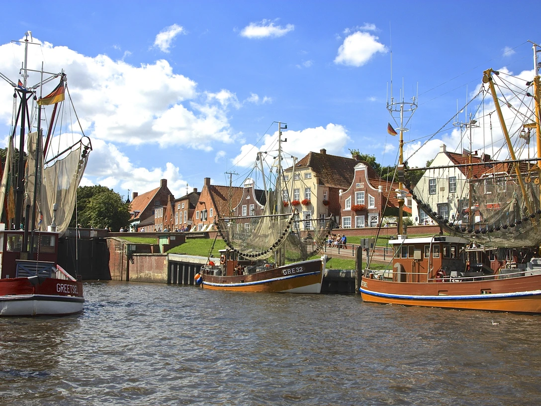 Hafen von Greetsiel Fischerboote mit hohen Masten im Hafen von Greetsiel, dahinter Backsteinhäuser unter blauem Himmel.