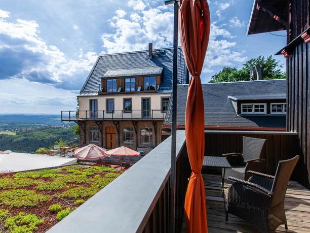 Uitzicht vanaf een terras met rotanmeubels op een groot gebouw met een leistenen dak, omgeven door een groen landschap en blauwe lucht.