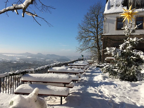 Aussicht an der Brand-Baude Verschneite Terrasse mit Holzbänken und Tischen, ein geschmückter Tannenbaum mit gelbem Stern, blauer Himmel und schneebedeckte Landschaft im Hintergrund.
