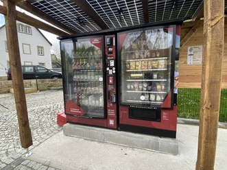 Proviantomat Standort in Schöna Roter Verkaufsautomat unter Solardach, gefüllt mit Getränken und Snacks, steht auf gepflastertem Platz neben einem Haus und Holzwand.