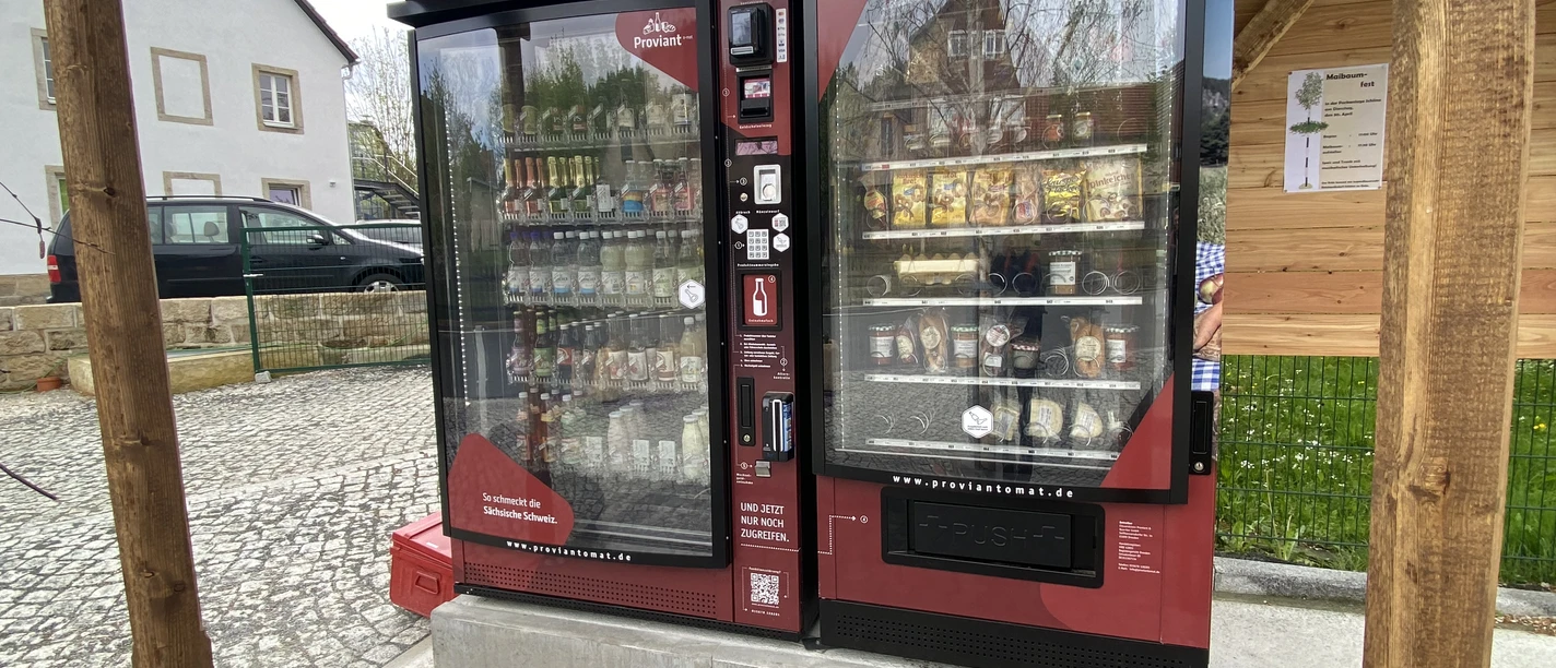Umístění Proviantomatu v Schöně Červený automat pod solární střechou, plný nápojů a občerstvení, stojí na dlážděném náměstí vedle domu a dřevěné zdi.