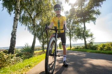 Rennrad Radfahrer auf einer sonnigen Landstraße, umgeben von Bäumen und Landschaft in Much, Nordrhein-Westfalen.