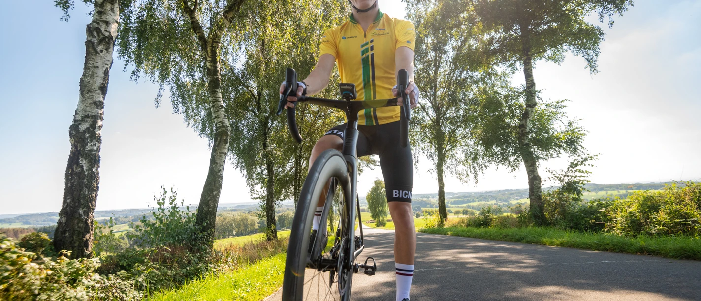 Rennrad Radfahrer auf einer sonnigen Landstraße, umgeben von Bäumen und Landschaft in Much, Nordrhein-Westfalen.