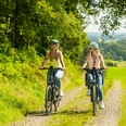Heckberg Zwei Frauen fahren bei sonnigem Wetter auf einem Feldweg durch eine grüne Landschaft mit Fahrrädern.