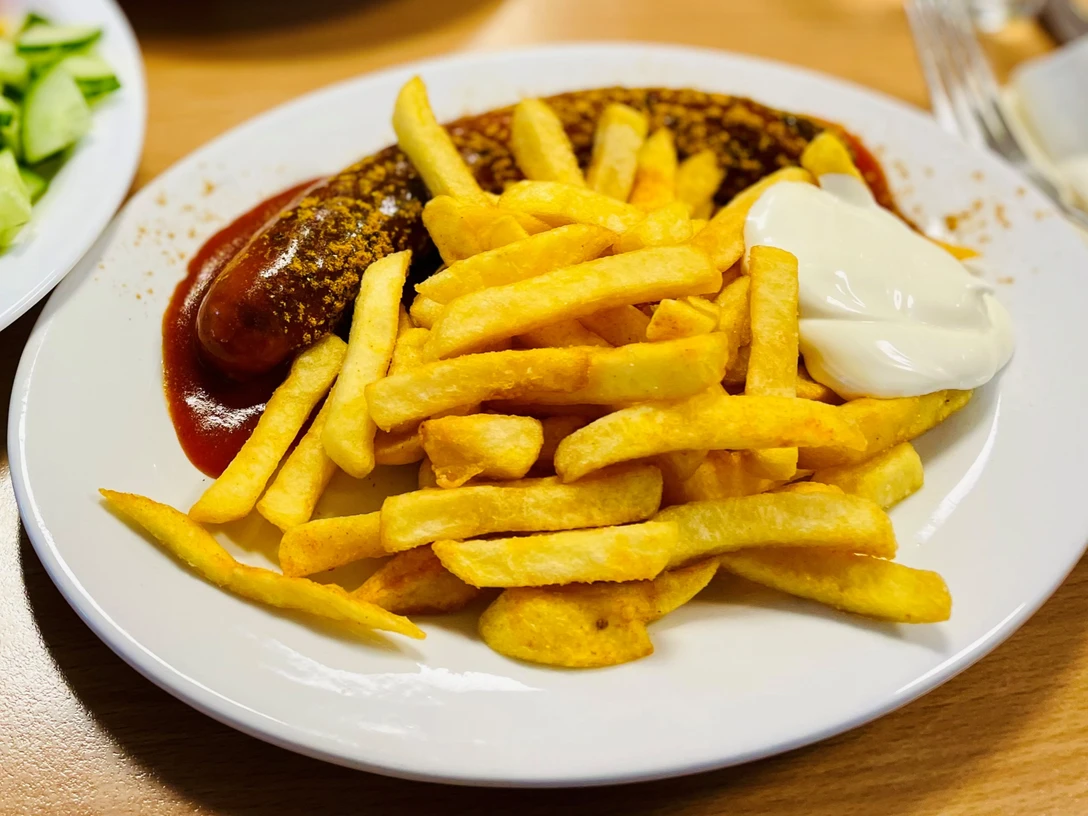 Speisen_GrillamMarkt_CC-BY-SA_Teutoburger_Wald_Stadt_Schloss_Holte-Stukenbrock@.jpg Currywurst mit Pommes frites und Mayonnaise auf einem weißen Teller, daneben ein Teller mit Gurkensalat.