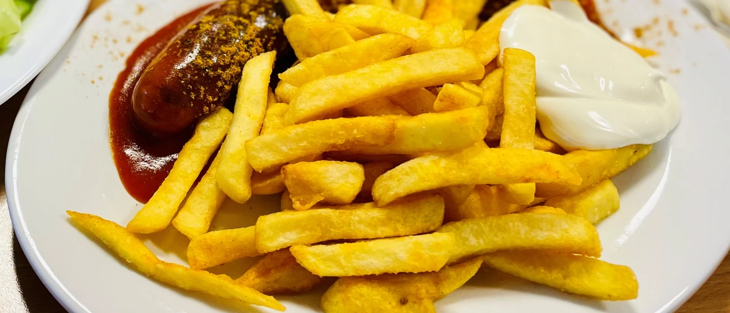 Speisen_GrillamMarkt_CC-BY-SA_Teutoburger_Wald_Stadt_Schloss_Holte-Stukenbrock@.jpg Currywurst mit Pommes frites und Mayonnaise auf einem weißen Teller, daneben ein Teller mit Gurkensalat.