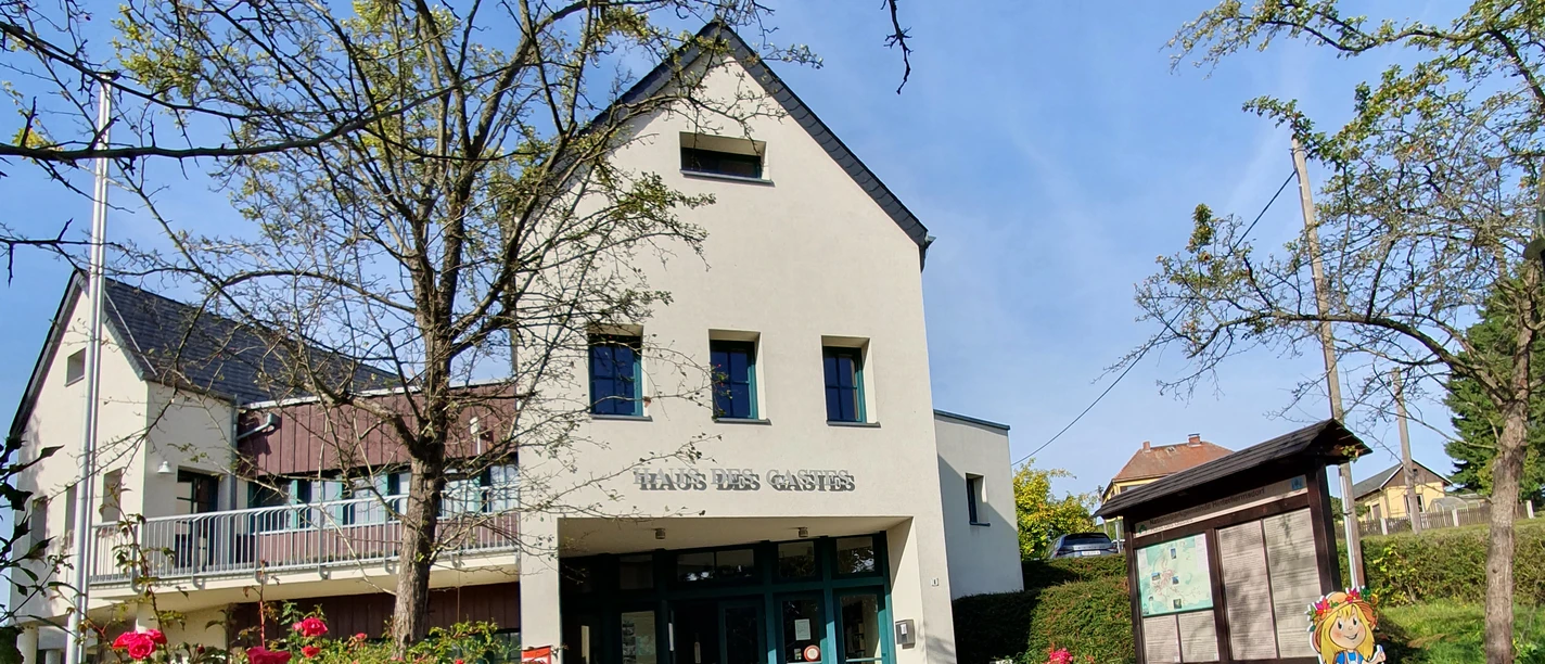 A bright building with the inscription "Haus des Gastes", surrounded by blooming roses and trees, under a clear blue sky.