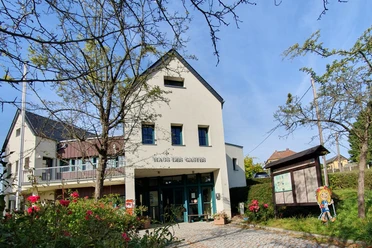 Haus des Gastes in Hinterhermsdorf Ein helles Gebäude mit der Aufschrift "Haus des Gastes", umgeben von blühenden Rosen und Bäumen, unter einem klaren blauen Himmel.