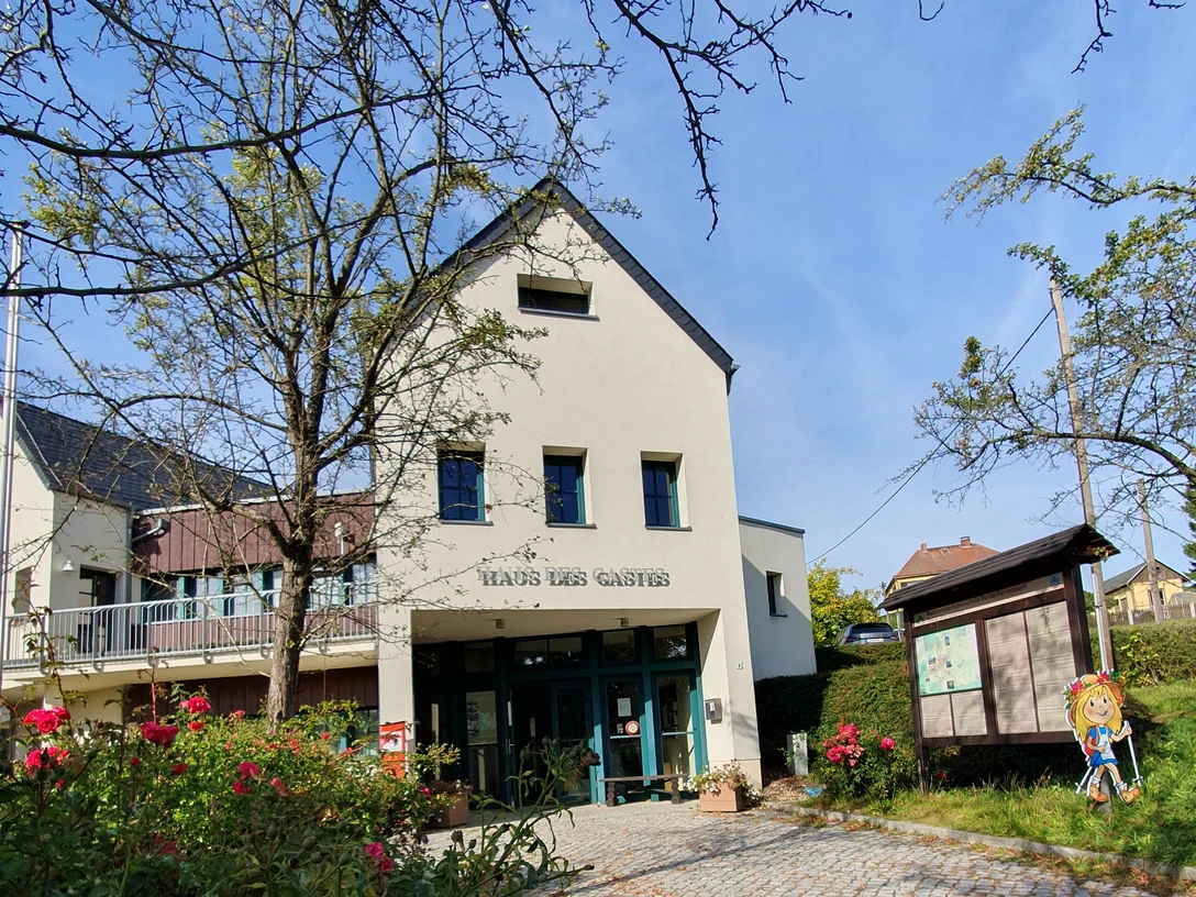 Haus des Gastes in Hinterhermsdorf Ein helles Gebäude mit der Aufschrift "Haus des Gastes", umgeben von blühenden Rosen und Bäumen, unter einem klaren blauen Himmel.
