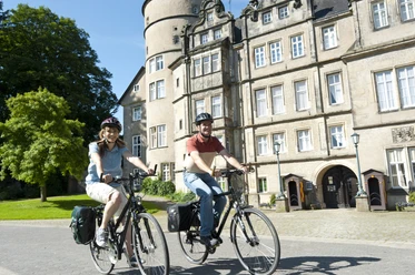 Schloss_Fahrrad2.jpg Zwei Radfahrer vor einem historischen Schloss mit Türmen und Zinnen radeln auf einem gepflasterten Weg.