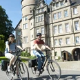 Schloss_Fahrrad2.jpg Zwei Radfahrer vor einem historischen Schloss mit Türmen und Zinnen radeln auf einem gepflasterten Weg.