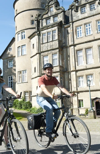 Schloss_Fahrrad2.jpg Zwei Radfahrer vor einem historischen Schloss mit Türmen und Zinnen radeln auf einem gepflasterten Weg.