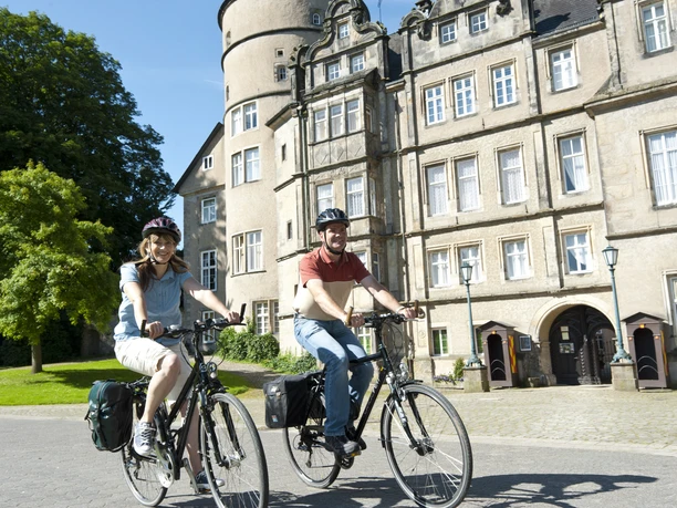 Schloss_Fahrrad2.jpg Zwei Radfahrer vor einem historischen Schloss mit Türmen und Zinnen radeln auf einem gepflasterten Weg.