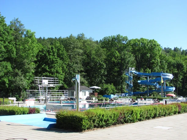 Venkovní bazén Berggießhübel / Copyright Stadt BGB.jpg Venkovní bazén Berggießhübel se skluzavkami a skokanskou plošinou, zasazený do zeleného lesního prostředí pod modrou oblohou.