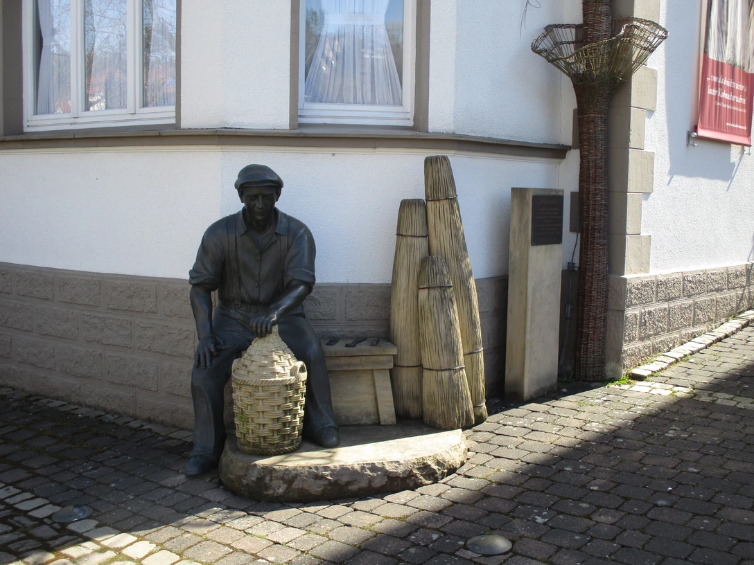 Korbmacher-Museum Dalhausen Eine Korbmacherstatue aus Bronze vor dem Museum, mit geflochtenen Körben und typischen Arbeitsgeräten.
