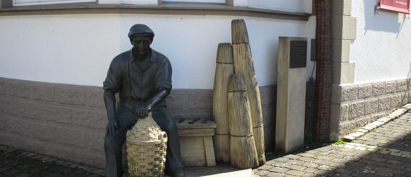 Korbmacher-Museum Dalhausen Eine Korbmacherstatue aus Bronze vor dem Museum, mit geflochtenen Körben und typischen Arbeitsgeräten.