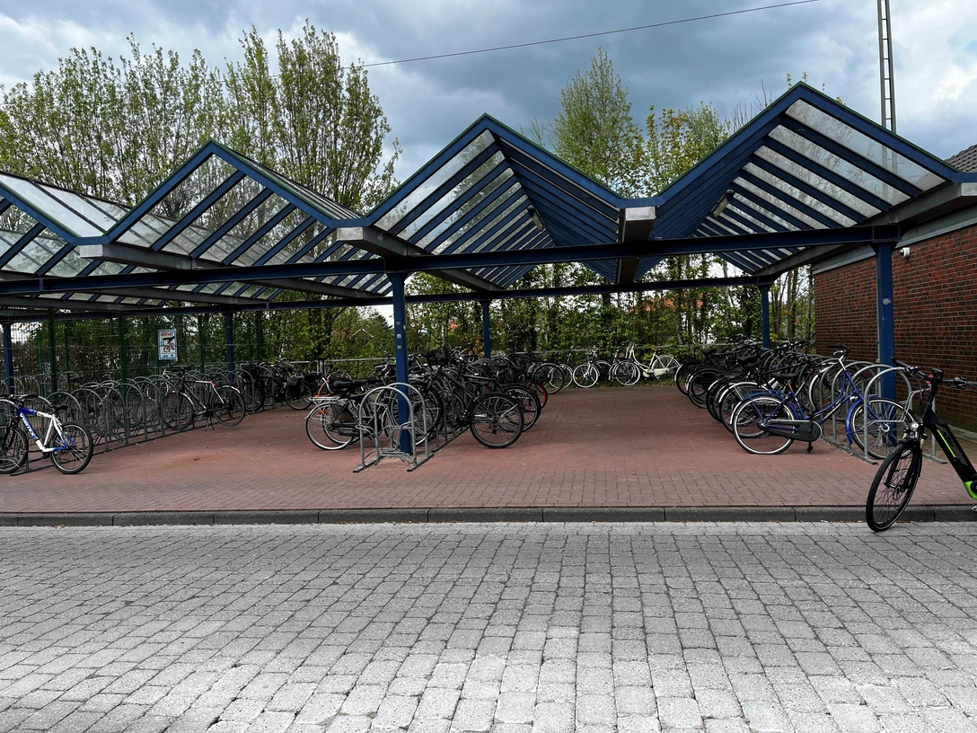 überdachte kostenlose Fahrradstellplätze am Bahnhof (Bahnhofstraße).jpg Überdachte Fahrradstellplätze aus Metall mit vielen geparkten Fahrrädern vor einem bewölkten Himmel.