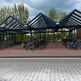 überdachte kostenlose Fahrradstellplätze am Bahnhof (Bahnhofstraße).jpg Überdachte Fahrradstellplätze aus Metall mit vielen geparkten Fahrrädern vor einem bewölkten Himmel.