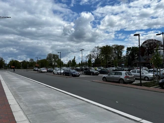 Parkplätze am Dockgelände.jpg Das Bild zeigt einen Parkplatz am Dockgelände an einem wolkigen Tag. Zahlreiche Autos sind auf dem asphaltierten Parkplatz geparkt, umgeben von ordentlich angelegten Gehwegen und Straßenlaternen. Im Hintergrund sind vereinzelte Bäume und Gebäude zu sehen, die eine urbane Umgebung andeuten.