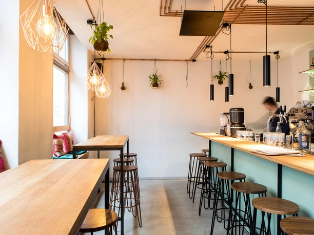 Tigermilch Interior view of a modern café featuring tall wooden tables and bar stools. Stylish pendant lights and greenery hang from the ceiling. A single person can be seen moving, adding a dynamic touch to the scene.