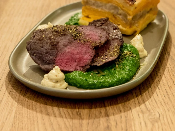 Tigermilch An elegantly presented dish arranged on an elongated plate: tender pink roasted meat accompanied by creamy green and white purées, alongside a savory, multilayered pastry glowing in orange hues and garnished with delicate vegetable crisps. This composition is placed on a natural wooden table.