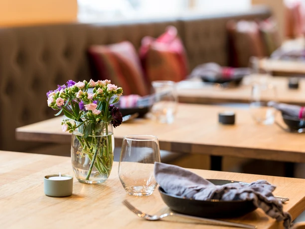 Tigermilch A stylishly arranged wooden table in a restaurant featuring colorful flowers in a glass vase, elegant glassware, and neatly folded napkins, creating an inviting atmosphere.