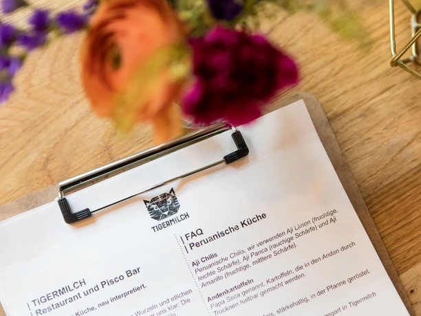 Tigermilch The image shows, in the foreground, a menu of the restaurant Tigermilch displayed on a clipboard resting on a wooden table, with blurred flowers in the background.