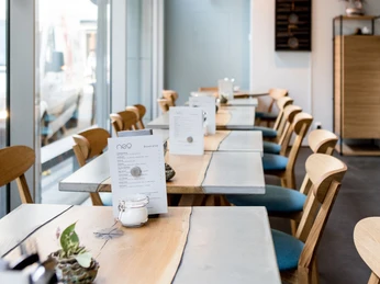 NeoBiota Helles, modernes Café mit Holzmöbeln und Menübroschüren auf jedem Tisch.Bright, modern café with wooden furniture and menu brochures on every table.