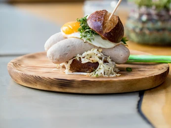 NeoBiota Ein kreativ angerichtetes Gericht auf einem runden Holzbrett: Zwei weiße Würste, garniert mit einem Spiegelei, frischen Kräutern und einem kleinen Laugenbrötchen, serviert auf einem Bett aus feinem Krautsalat. Ein Holzstäbchen hält die Köstlichkeit zusammen, während im Hintergrund unscharfe Pflanzen und helle Tische sichtbar sind.A creatively arranged dish on a round wooden board: two white sausages garnished with a fried egg, fresh herbs and a small pretzel roll, served on a bed of fine coleslaw. A wooden skewer holds the delicacy together, while blurred plants and light-coloured tables are visible in the background.