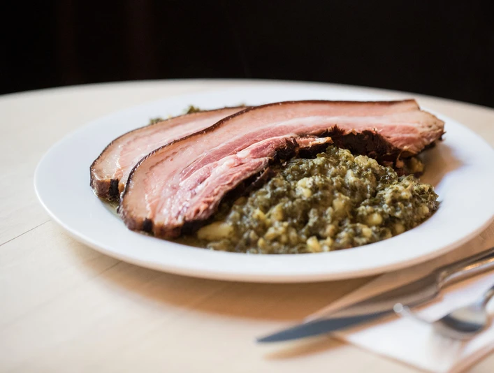 Max Stark Ein Teller mit zwei Scheiben geräuchertem Fleisch, serviert auf einem Bett aus Grünkohl und Kartoffeln.A plate with two slices of smoked meat served on a bed of kale and potatoes.
